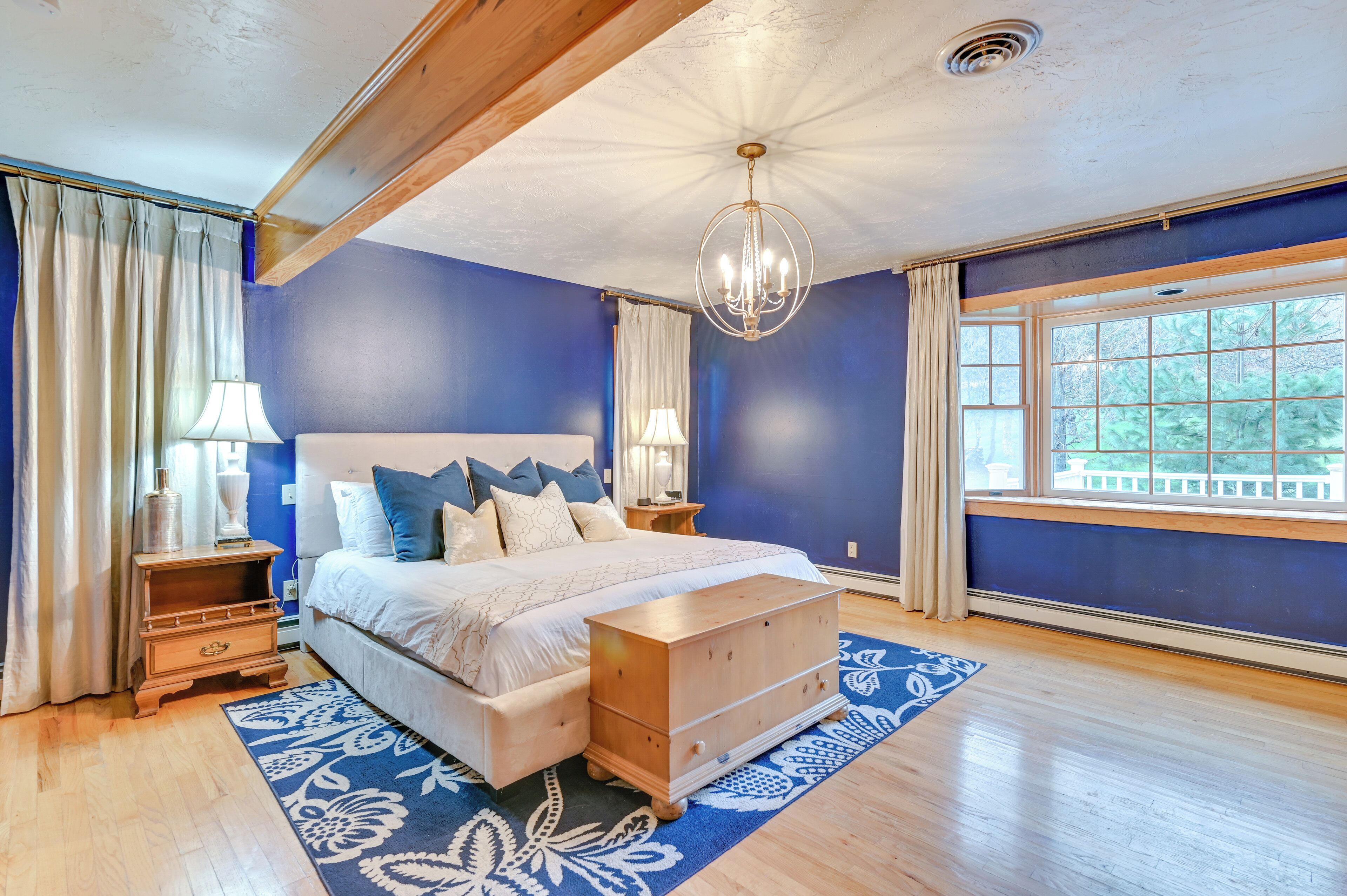 A bedroom with a large bed, wooden headboard, a chandelier, a nightstand, a dresser, and a window with a view of trees.