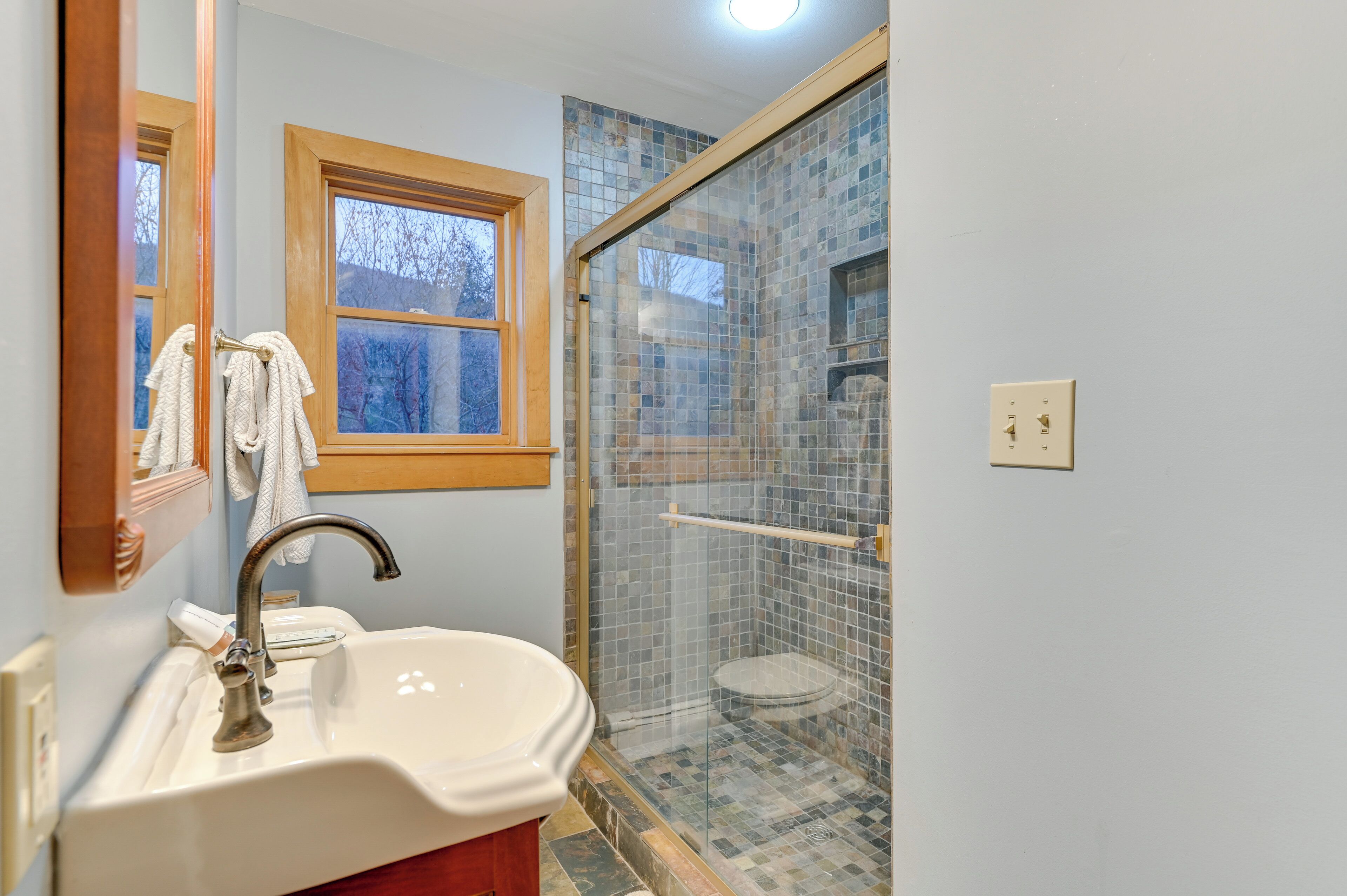 A bathroom with a sink, mirror, window, and a glass-enclosed shower area.
