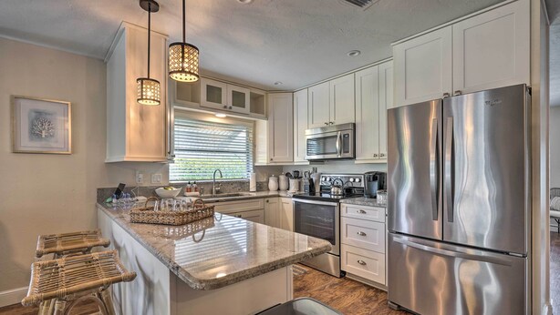A modern kitchen with white cabinets, stainless steel appliances, a granite countertop, wicker bar stools, and pendant lighting. - Naples Oasis w/ Screened Pool, Bike to Beach! (Naples)