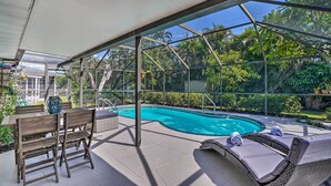 A covered outdoor pool area with a glass enclosure, wooden furniture, and a view of lush greenery. - Naples Oasis w/ Screened Pool, Bike to Beach! (Naples)