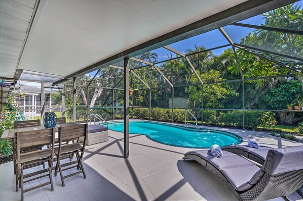 An outdoor pool area with a covered patio, seating, and lush greenery.