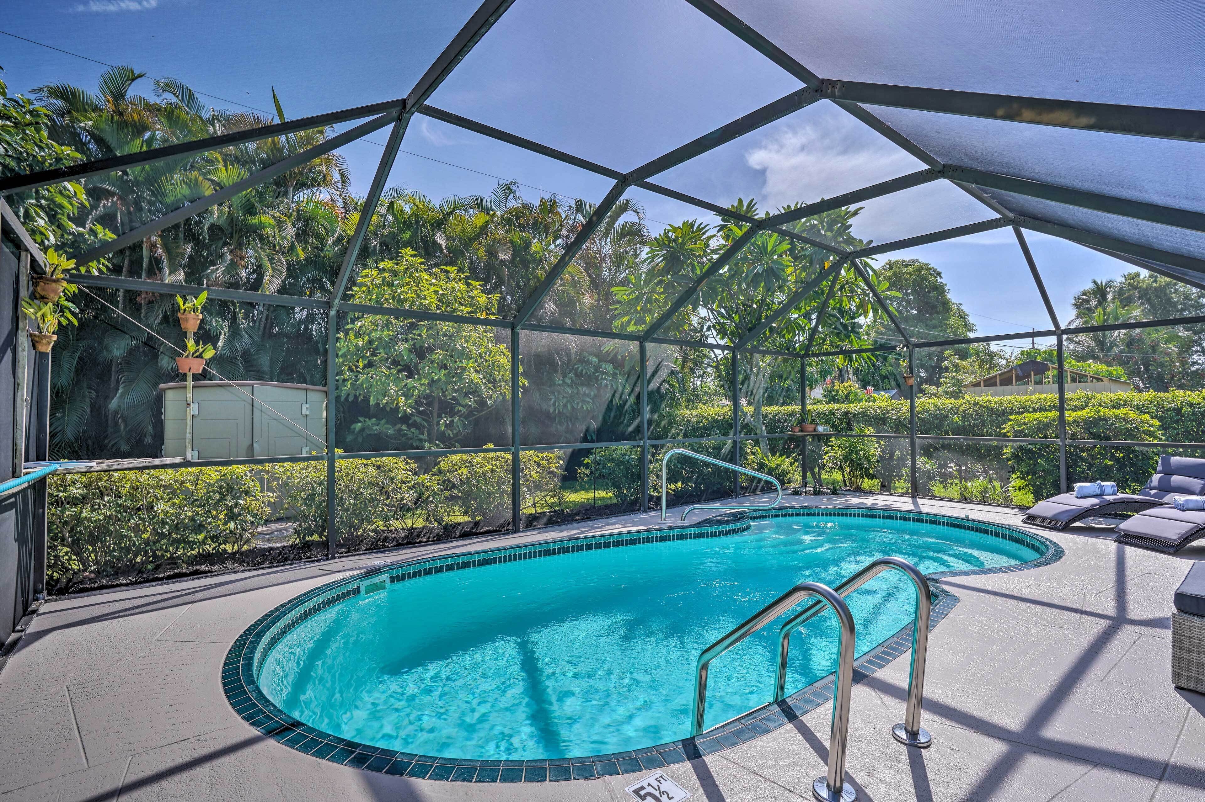 A covered outdoor swimming pool with a glass enclosure, surrounded by lush greenery and a clear blue sky.