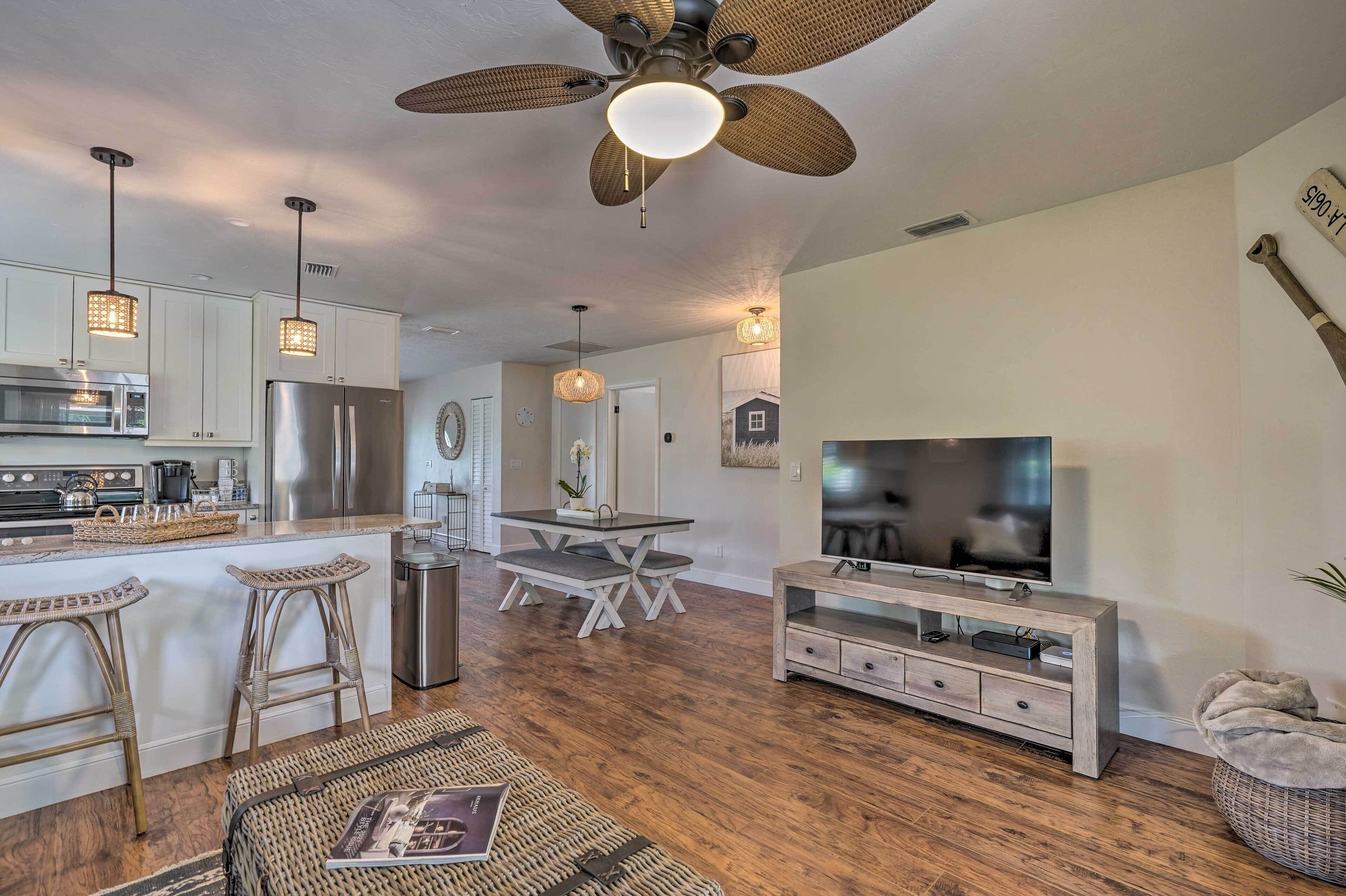 A modern kitchen with stainless steel appliances, a dining area with a table and chairs, a TV on a wooden stand, and a ceiling fan.