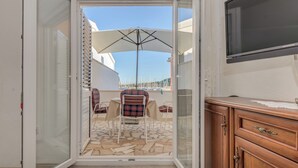Een kamer met een schuifglazen deur die uitkomt op een terras met een tafel, stoelen en een grote parasol. Aan de muur hangt een televisie.
