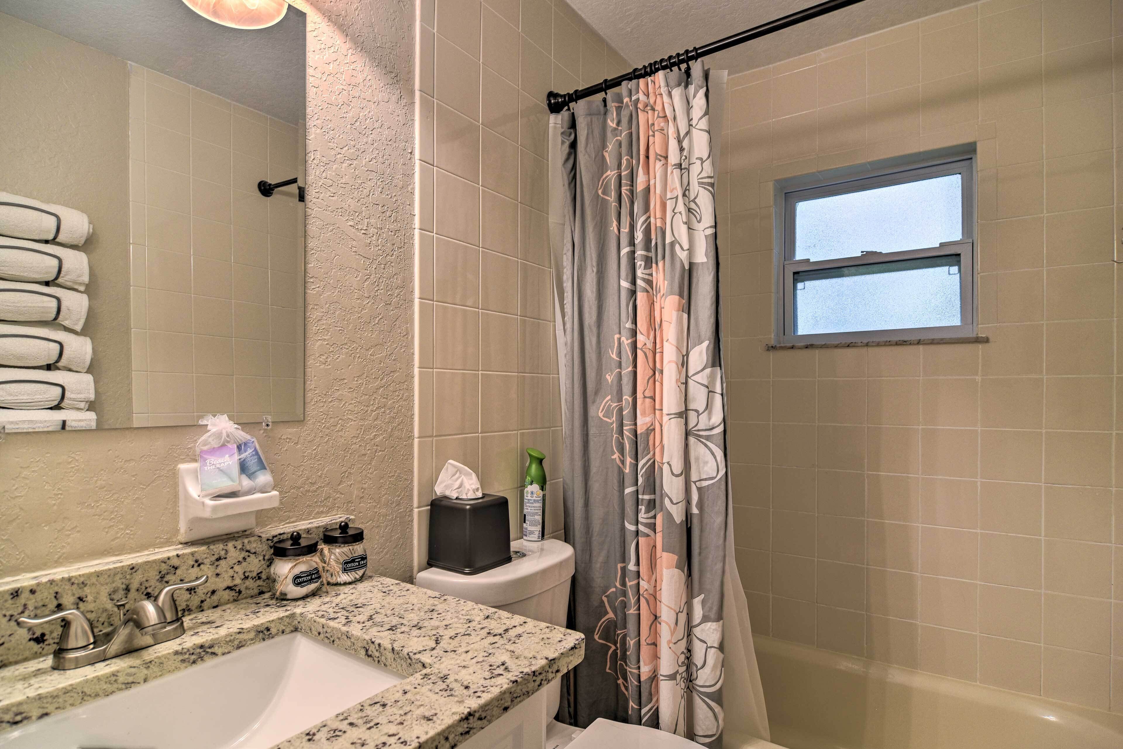 A bathroom with a granite countertop sink, a toilet with a tissue box and a bottle, a shower curtain with floral patterns, and a window above the toilet.