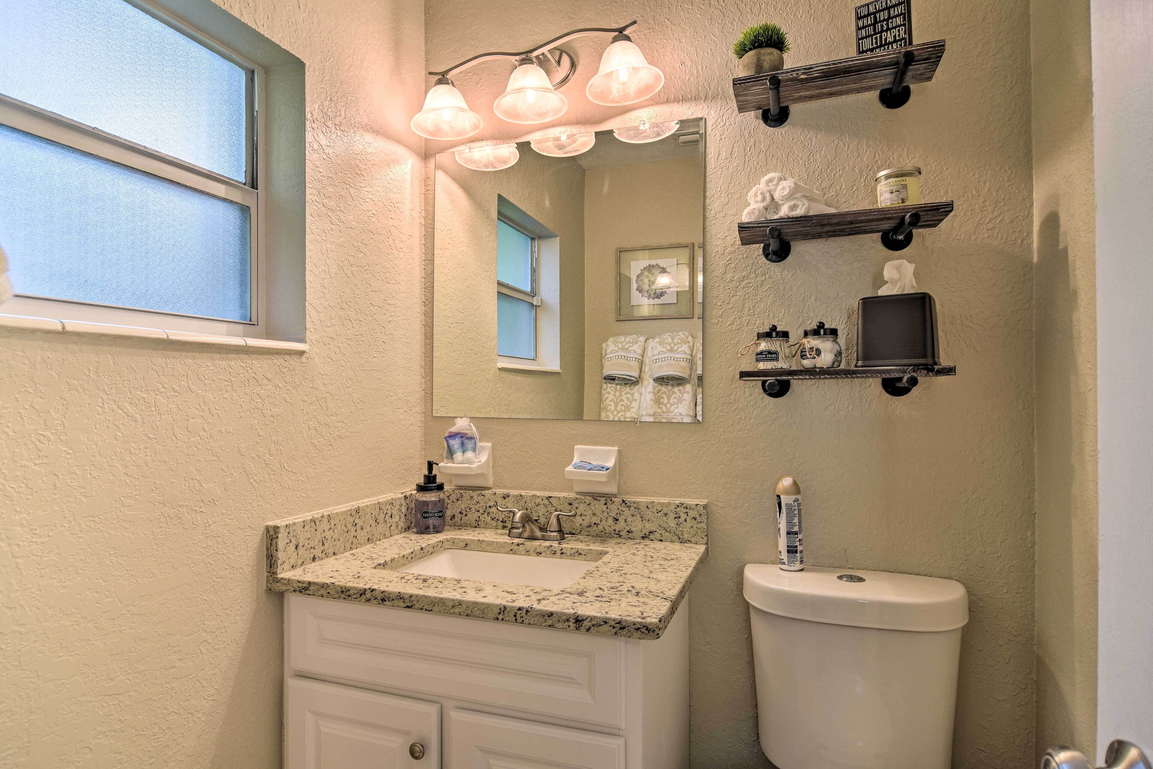 Une salle de bain avec un lavabo surmonté d’un plan de travail en granit, des meubles blancs, des toilettes et des étagères murales où sont rangés les articles de toilette.