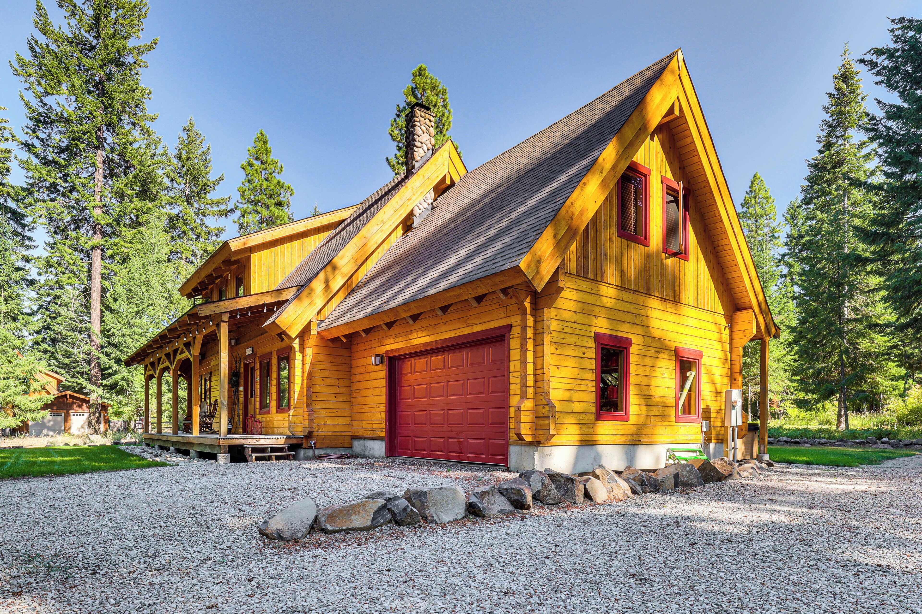 A yellow wooden cabin with a red garage door, surrounded by a gravel driveway and a stone wall.