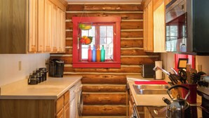 A log cabin kitchen with wooden cabinets, a window with colorful bottles, a sink, and a kettle. - Award-winning Log Cabin, Top 5 in New England! (Londonderry)