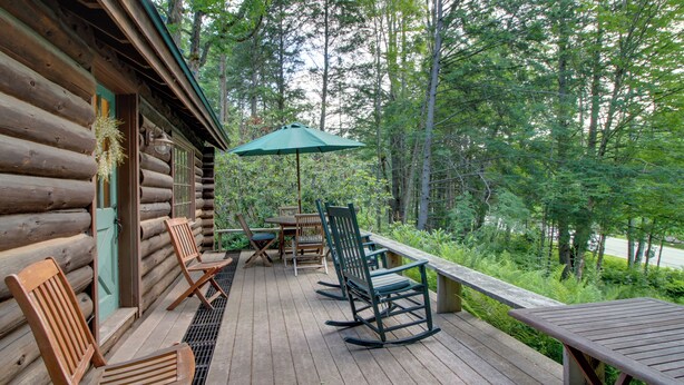 A wooden deck with outdoor furniture, including chairs and a table, overlooking a forested area. - Award-winning Log Cabin, Top 5 in New England! (Londonderry)