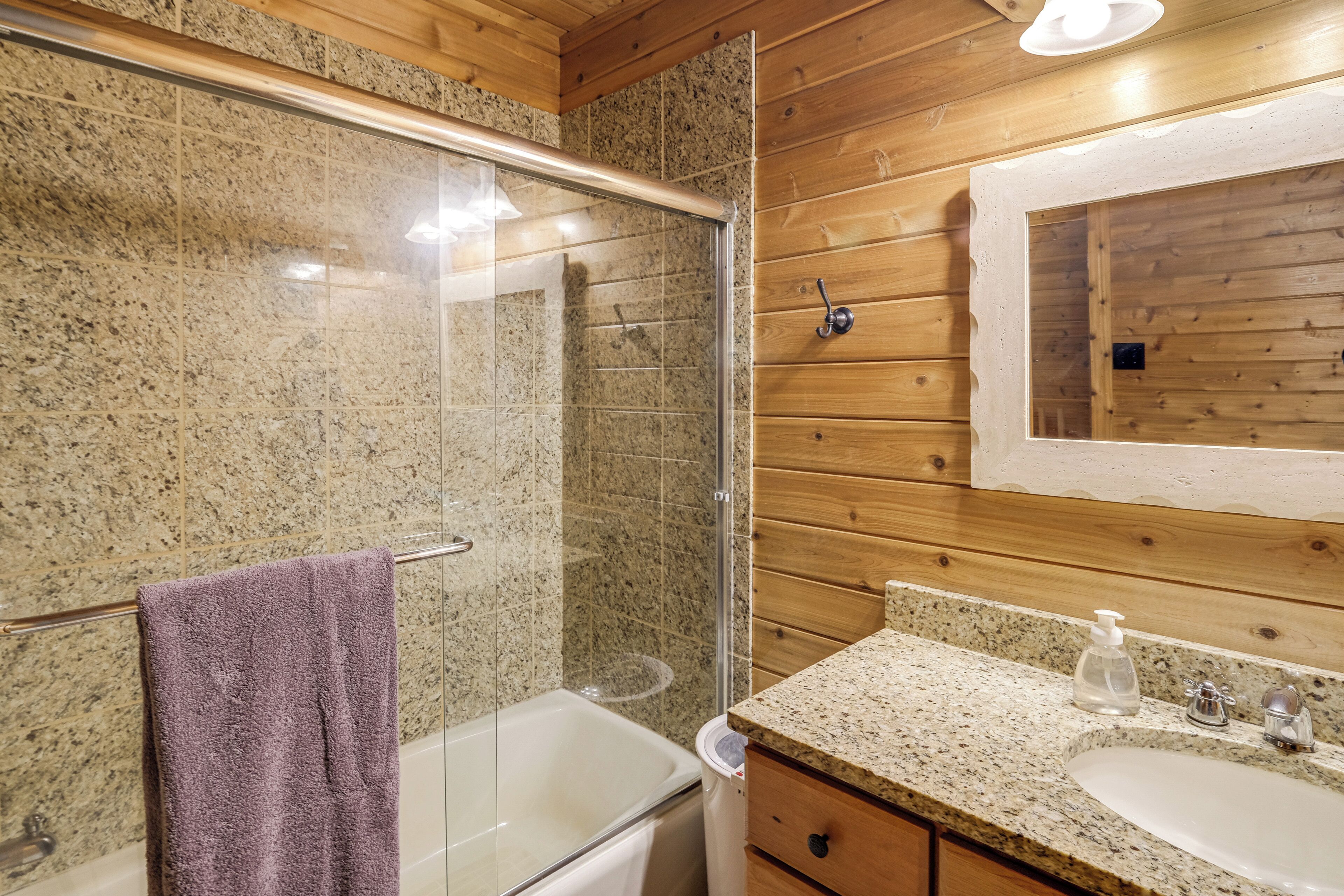 A bathroom with a glass-enclosed shower, a white bathtub, a granite countertop, and wooden paneling.