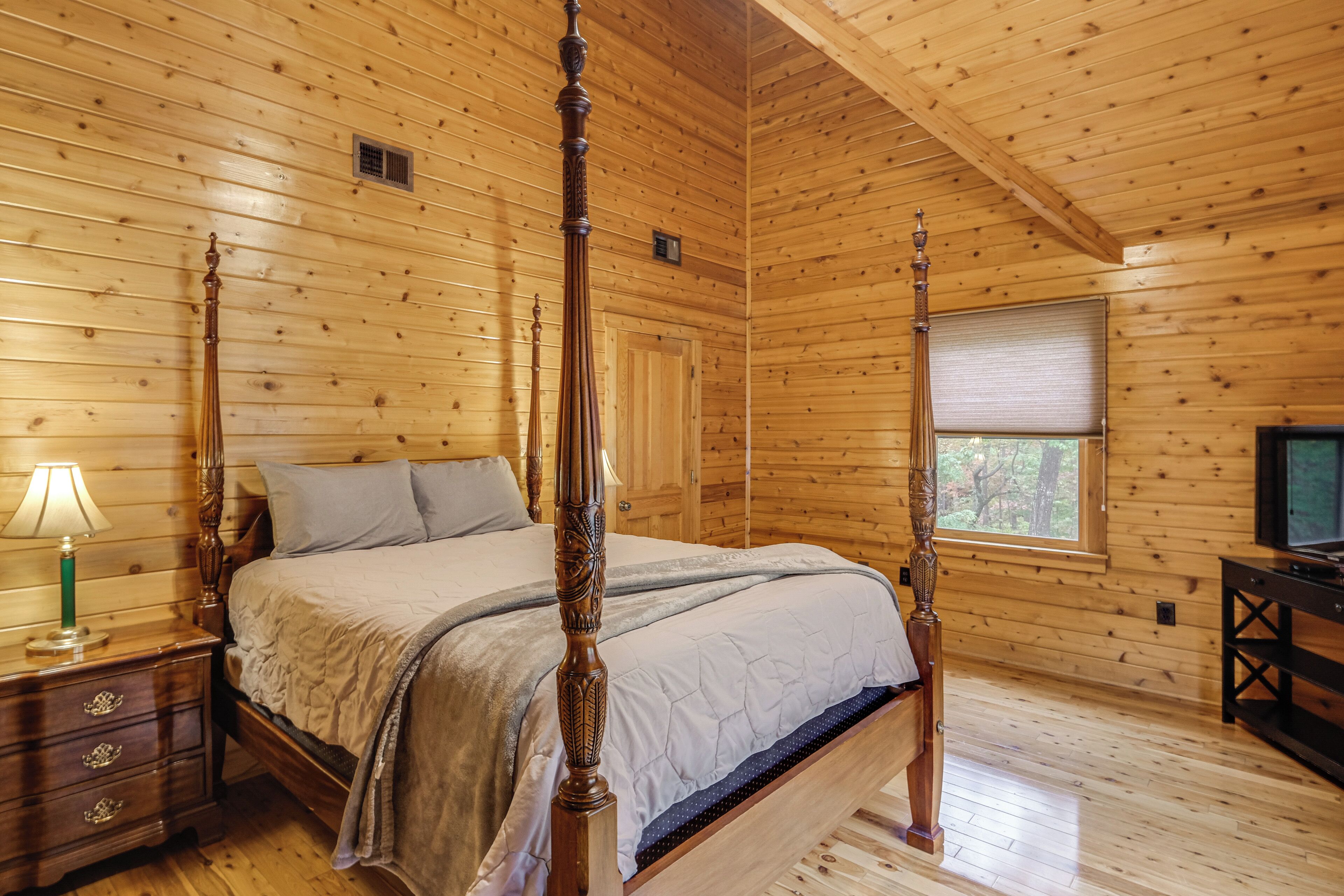 A four-poster bed in a wooden cabin room with a window and a television on a wooden stand.