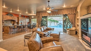 A spacious living room with a leather sectional sofa, a flat-screen TV, and a large window offering a view of a pool and palm trees. - Heated Pool + Putting Green: Luxe Gilbert Home (Gilbert)