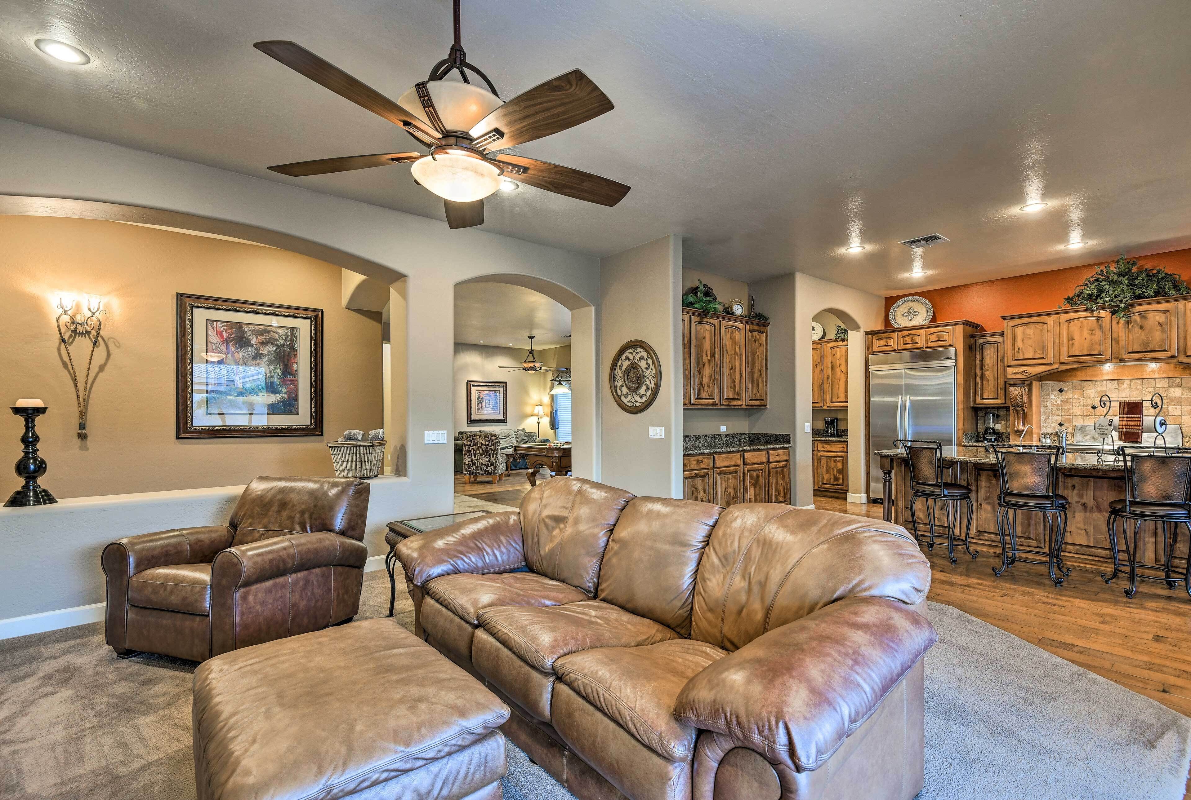 A spacious living room with a leather sofa, a ceiling fan, and a kitchen area with wooden cabinets and a stainless steel refrigerator.