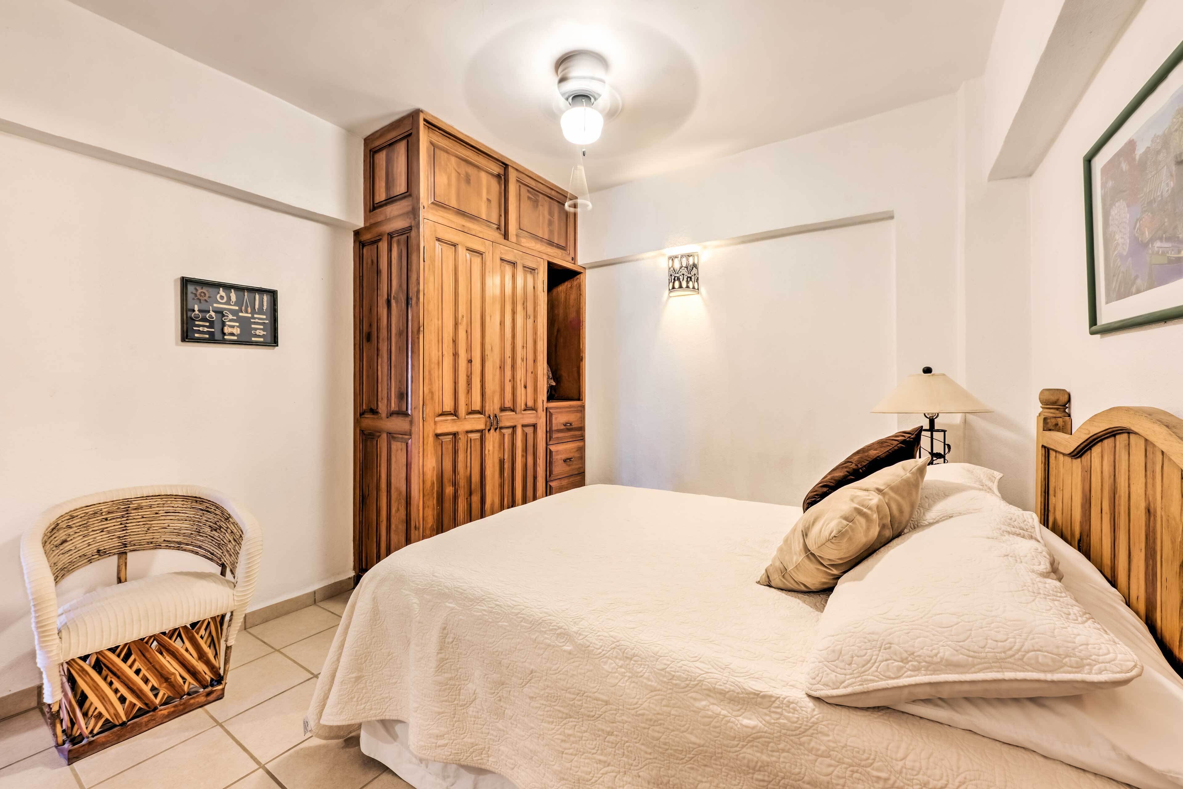 A bedroom with a bed, wooden wardrobe, wicker chair, and a wall-mounted light fixture.
