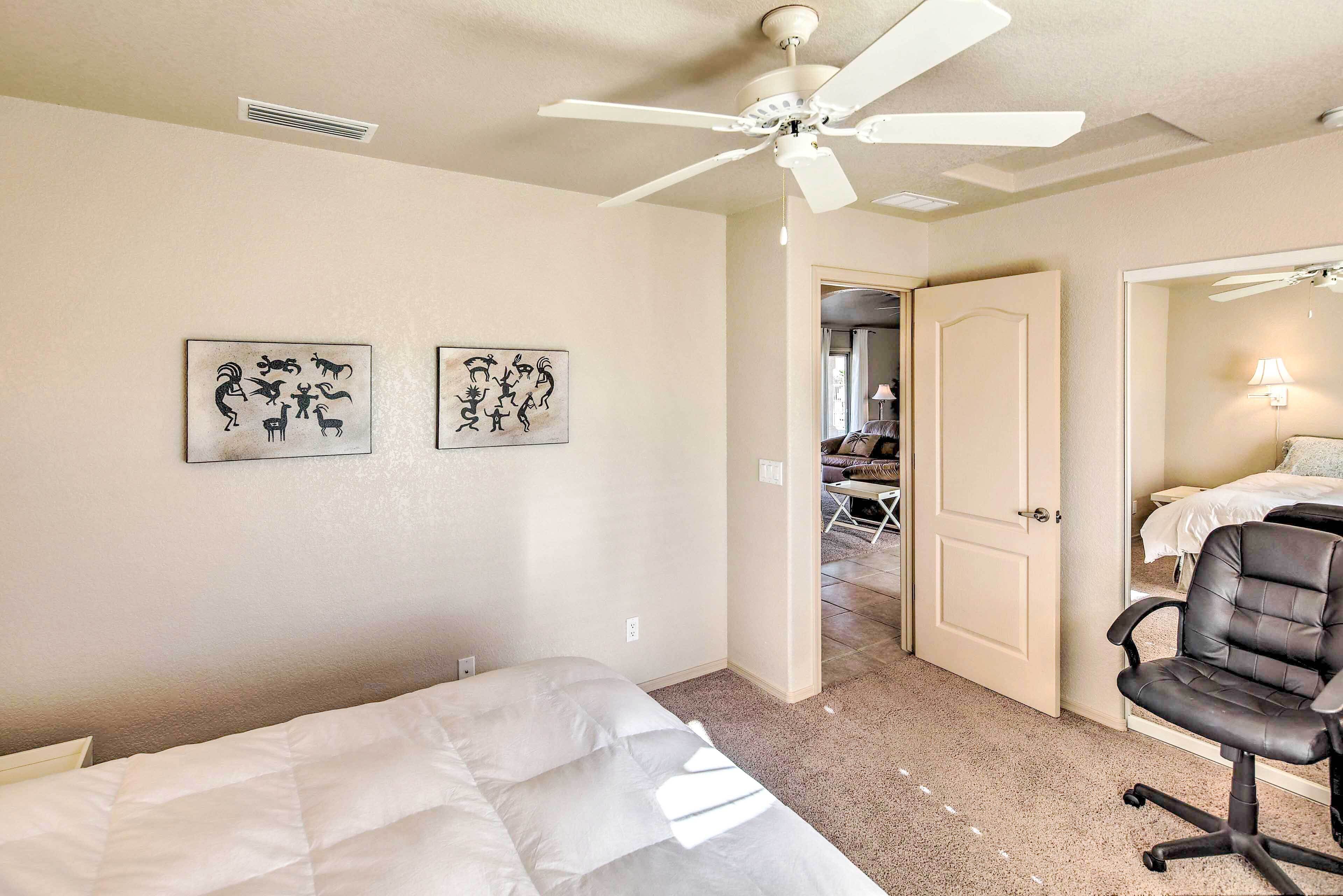 A hotel room with a bed, a desk, a chair, a ceiling fan, and two wall art pieces.