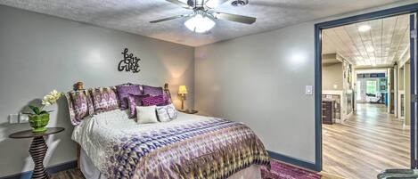 Un dormitorio con cama, ventilador de techo, mesita de noche, lámpara y vista al pasillo.