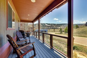 Um deck de madeira com duas cadeiras Adirondack, uma grade de metal e vista para uma paisagem montanhosa com uma estrada e edifícios ao longe.