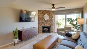 A living room with a stone fireplace, a flat-screen TV, a sofa, and a ceiling fan. - First-floor Condo w/ Golf Course Views & Pool! (Scottsdale)