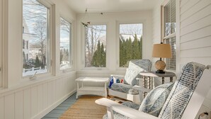 A cozy room with a large window, a wicker chair, a patterned armchair, and a small table with a lamp. - < 1 Mi to Beach! Kennebunk Cottage w/ Yard (Kennebunk)
