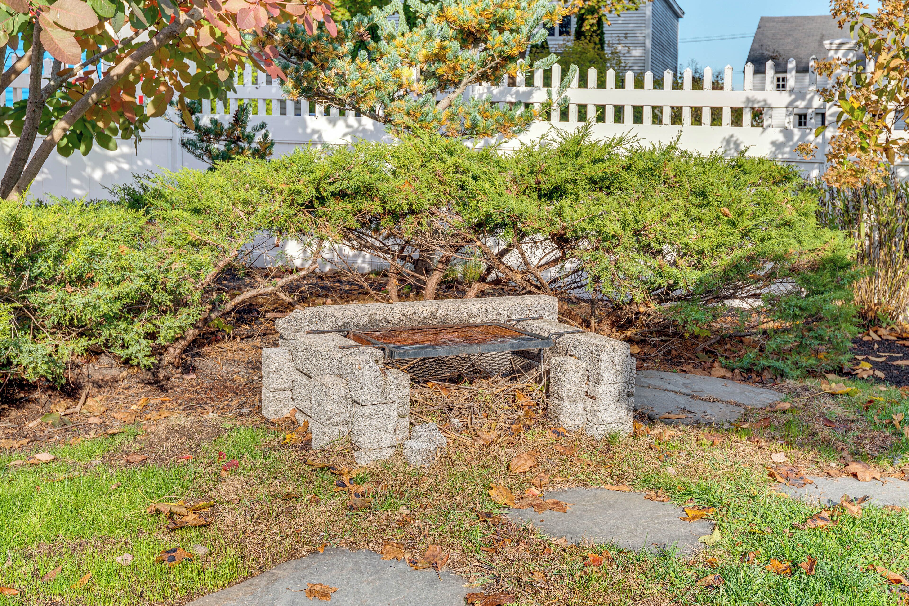 A stone bench surrounded by greenery, with a white picket fence and a building in the background.