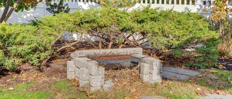A stone bench surrounded by greenery, with a white picket fence and a building in the background.