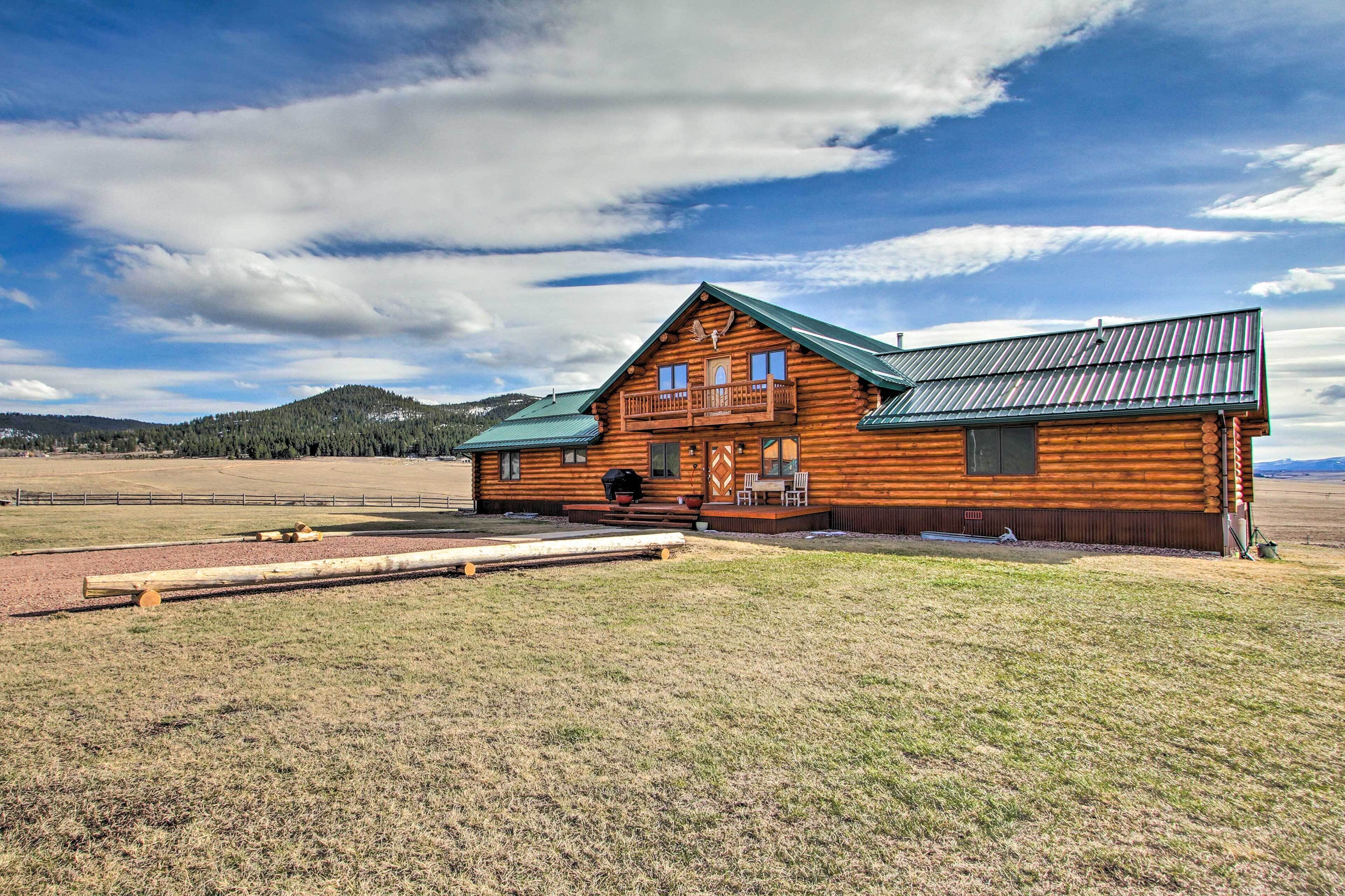 A log cabin with a balcony, a green roof, and a view of a field and mountains in the background.