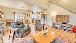 A spacious living room with a leather sofa, a patterned armchair, and a wooden coffee table. A kitchen is visible in the background with a refrigerator and a dining area. - Family-friendly Bend Home w/ Hot Tub & Yard! (Bend)