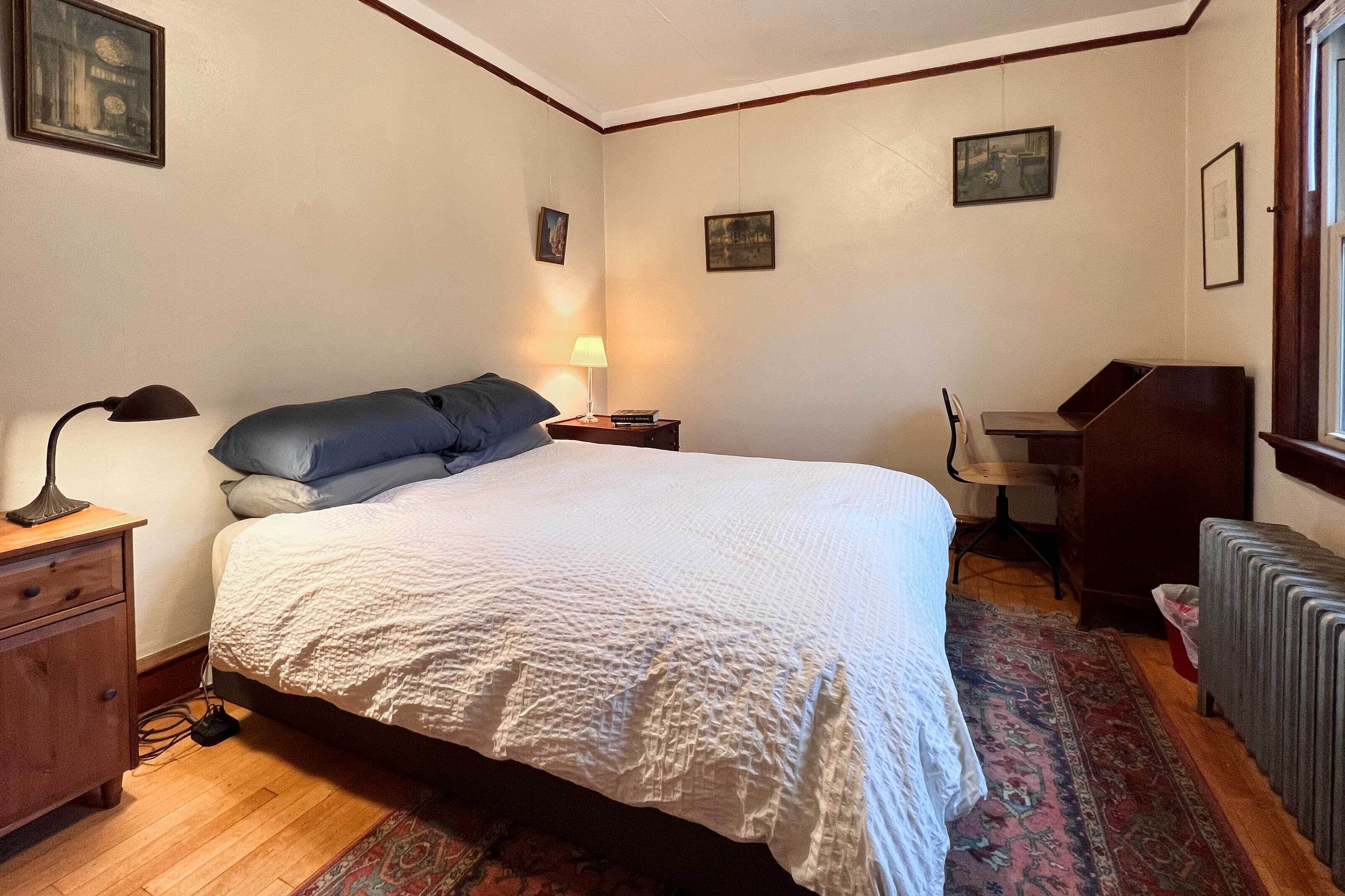 A bedroom with a large bed, a desk, a chair, a radiator, and a nightstand with a lamp.