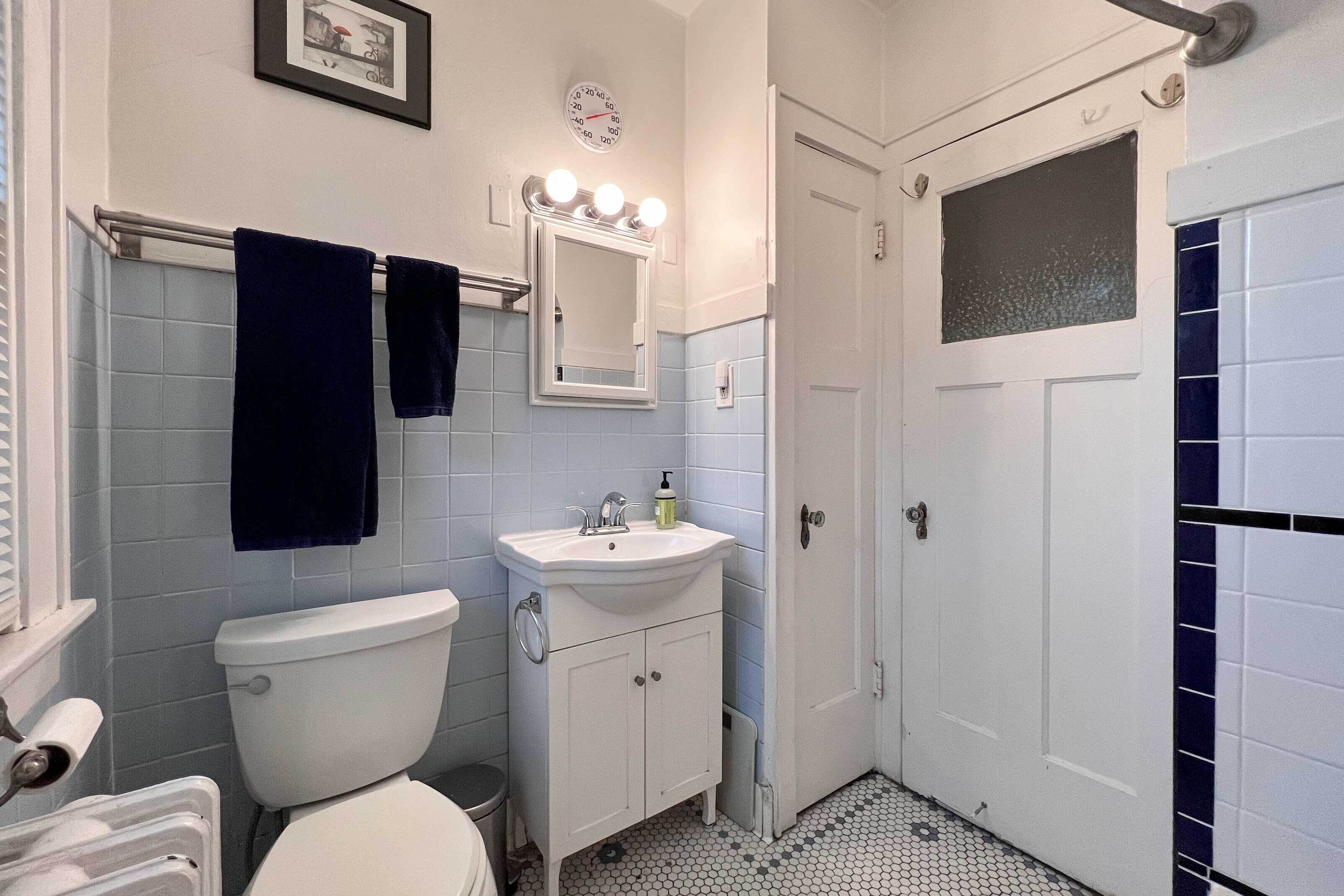 Un baño con inodoro, lavabo y espejo. Hay un perchero con dos toallas, un reloj en la pared y una ventana con puerta de vidrio esmerilado.