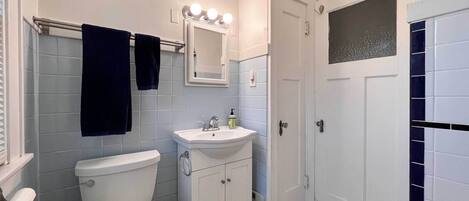 Un baño con inodoro, lavabo y espejo. Hay un perchero con dos toallas, un reloj en la pared y una ventana con puerta de vidrio esmerilado.