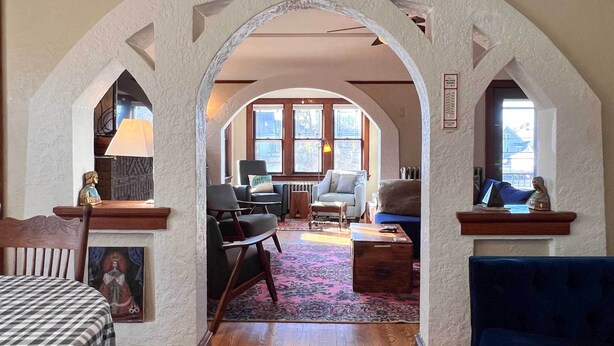 A cozy living room with a large arched doorway, wooden furniture, and a view through the windows.