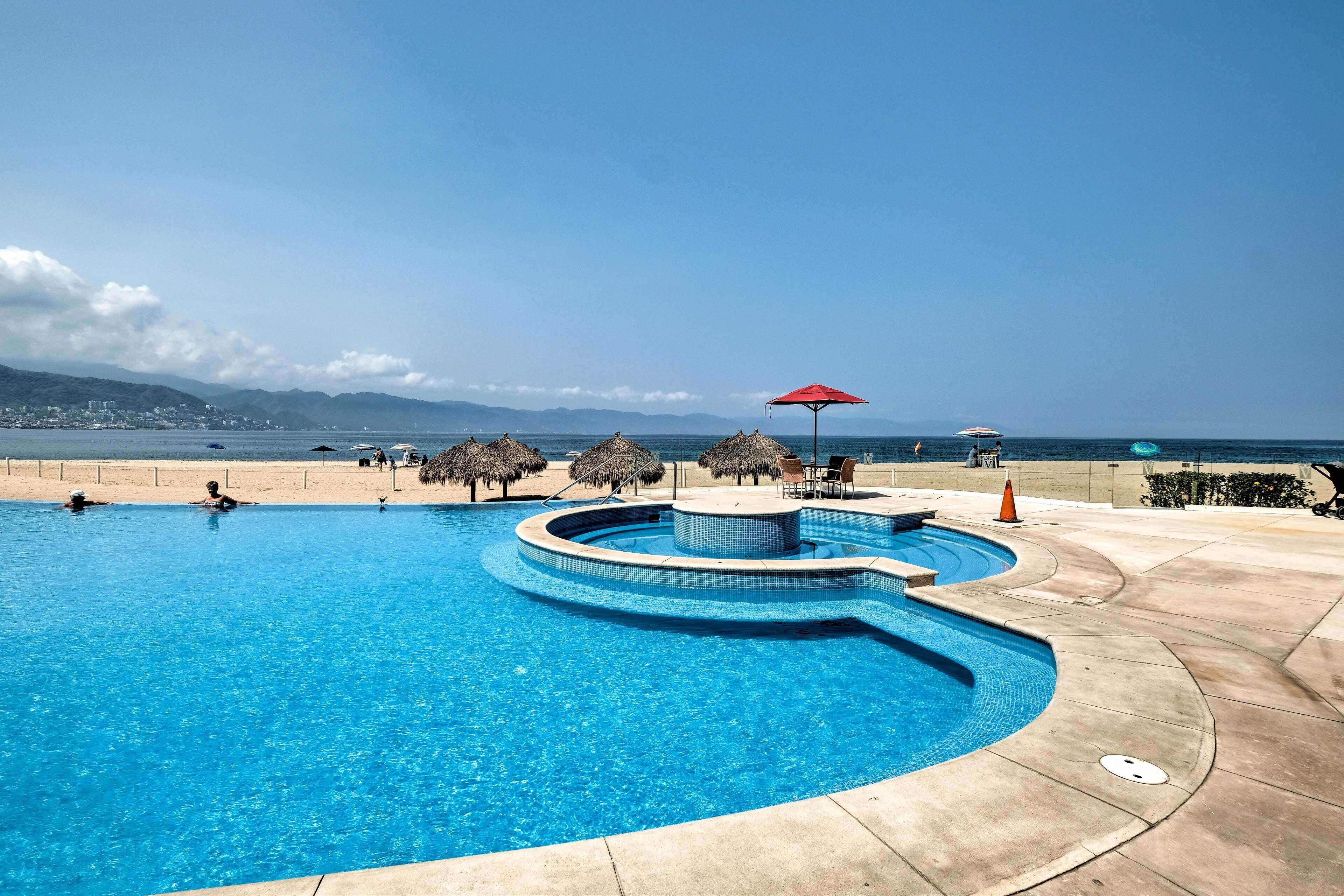A swimming pool with a curved design, surrounded by a concrete deck and thatched umbrellas.