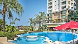 A swimming pool with a circular design, surrounded by palm trees and a multi-story building in the background. - Beachfront Resort Condo w/ Wraparound Balcony! (Puerto Vallarta)