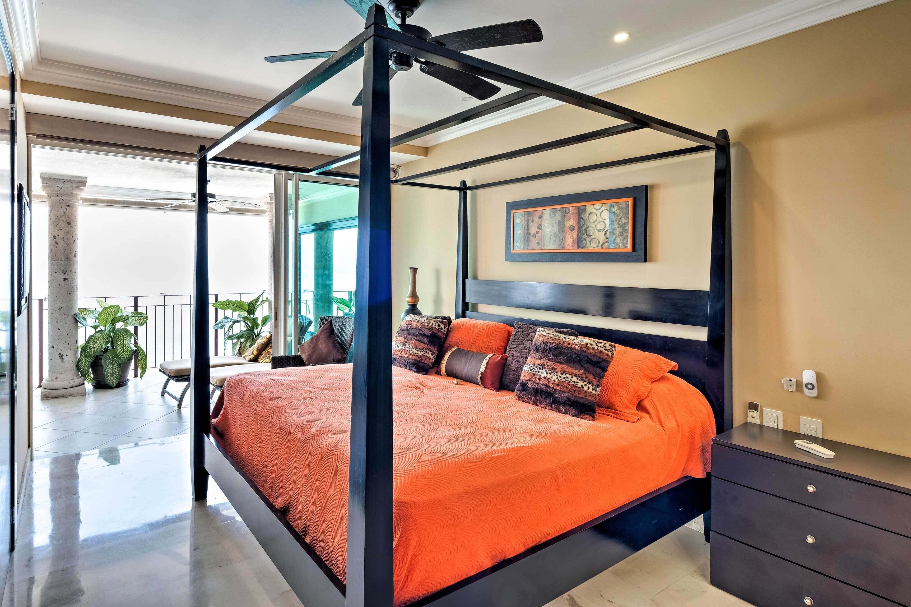 A four-poster bed with an orange bedspread, a dark wooden headboard, and multiple decorative pillows. There is a nightstand with a remote control and a wall-mounted light switch.