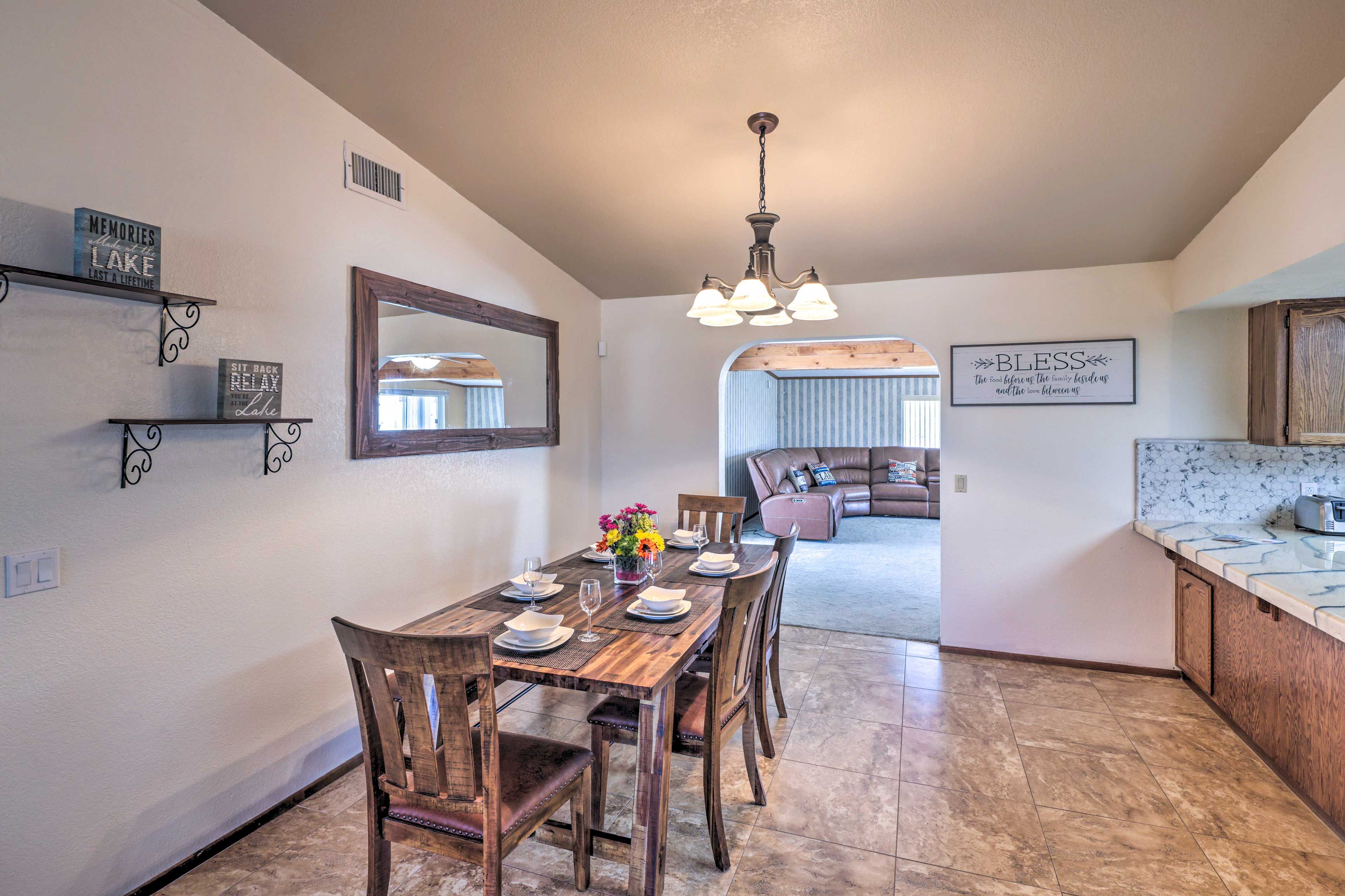 Un comedor con mesa de madera dispuesta para seis personas, una araña y una cocina con encimera de mármol.