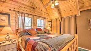 Un dormitorio de cabaña de madera con una cama grande, una mesita de noche y un ventanal con cortinas.