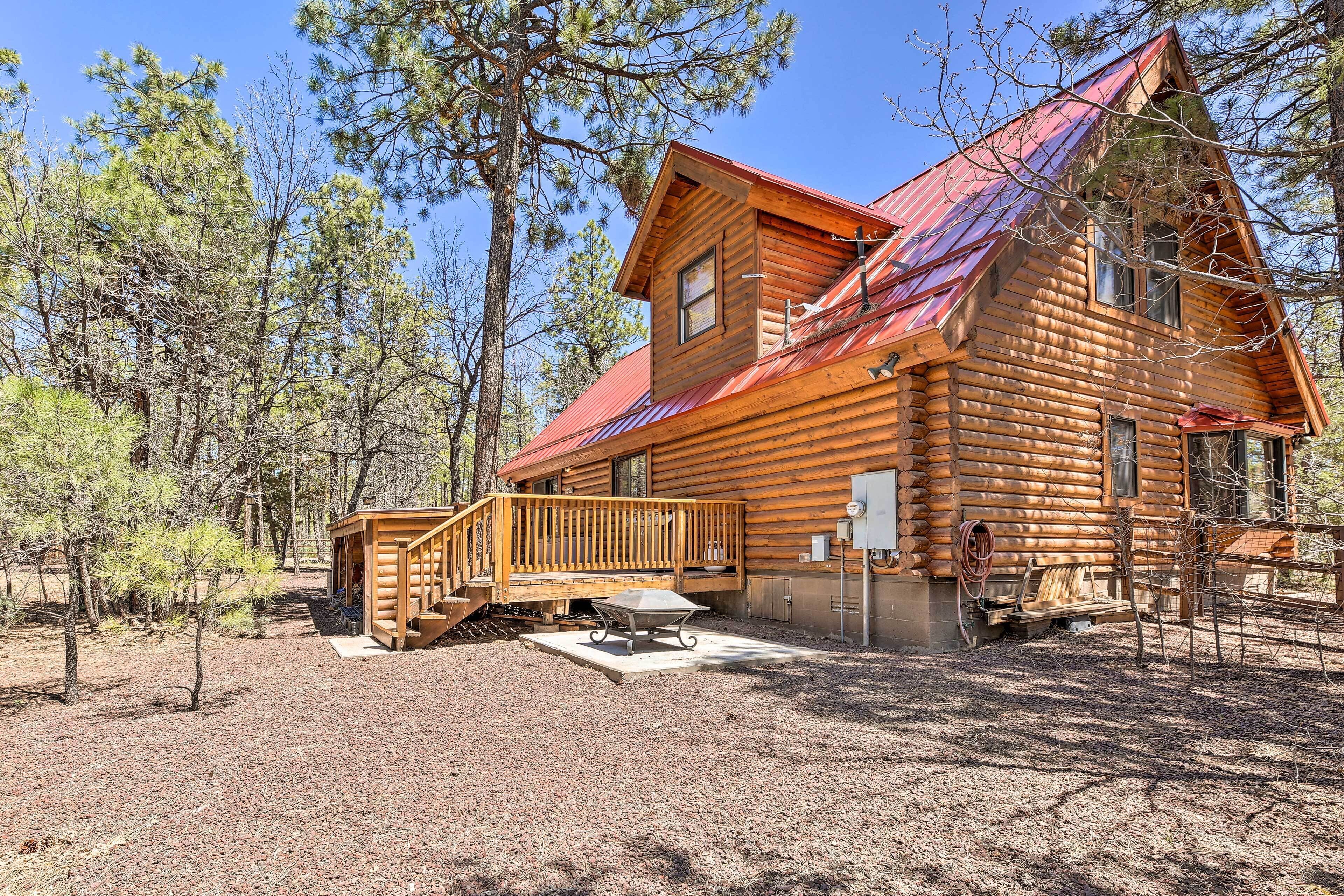 A log cabin with a red metal roof, a wooden deck, and a picnic table in a forested area.