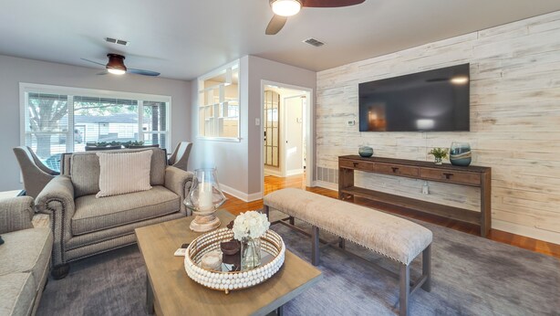 A living room with a sofa, coffee table, and bench, a ceiling fan, and a TV mounted on the wall. - Lovely Lubbock Home w/ Furnished Deck & Grill (Lubbock)