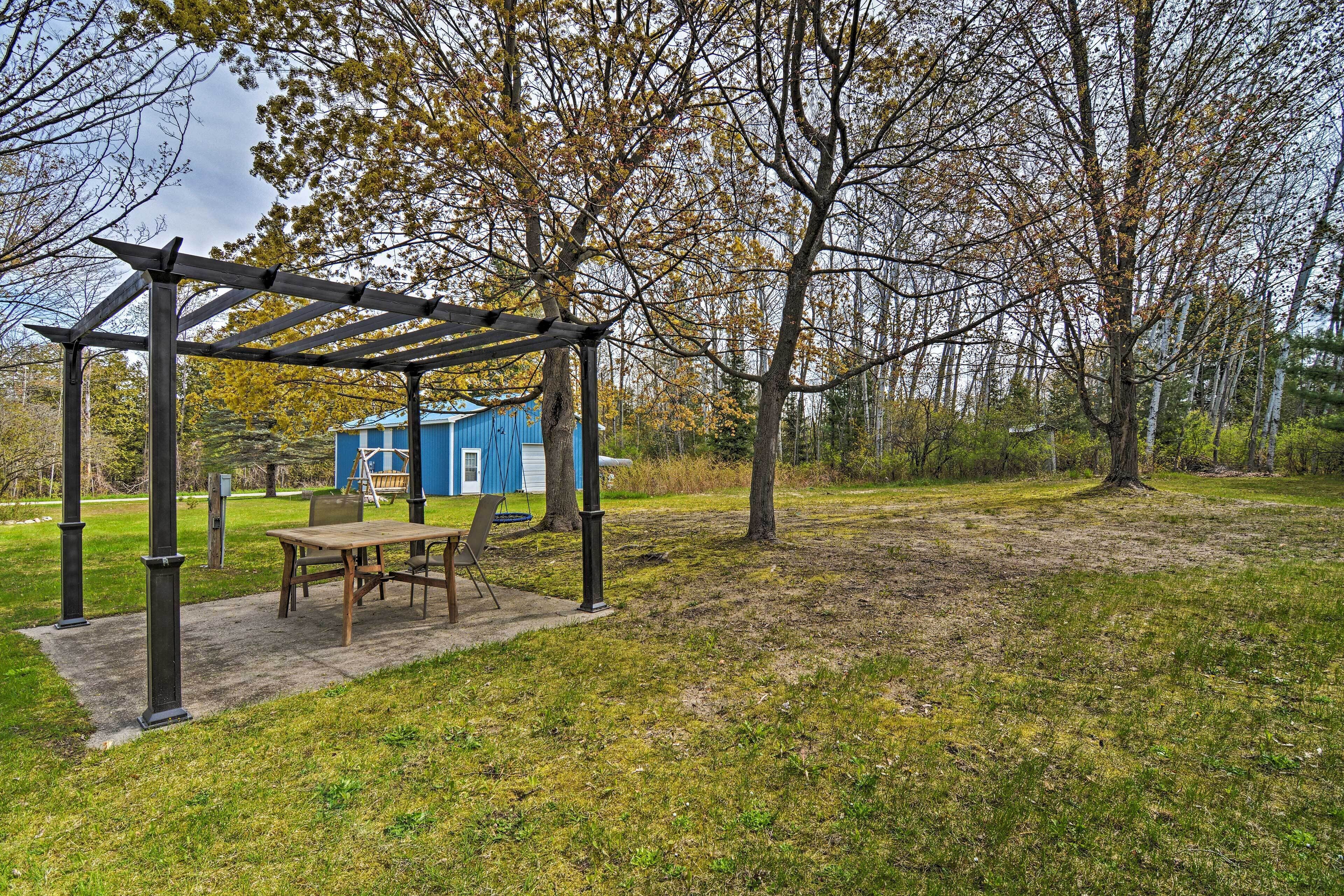 Una pérgola de madera con una mesa y sillas, un cobertizo azul y un área con césped y árboles.