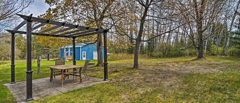 Una pérgola de madera con una mesa y sillas, un cobertizo azul y un área con césped y árboles.