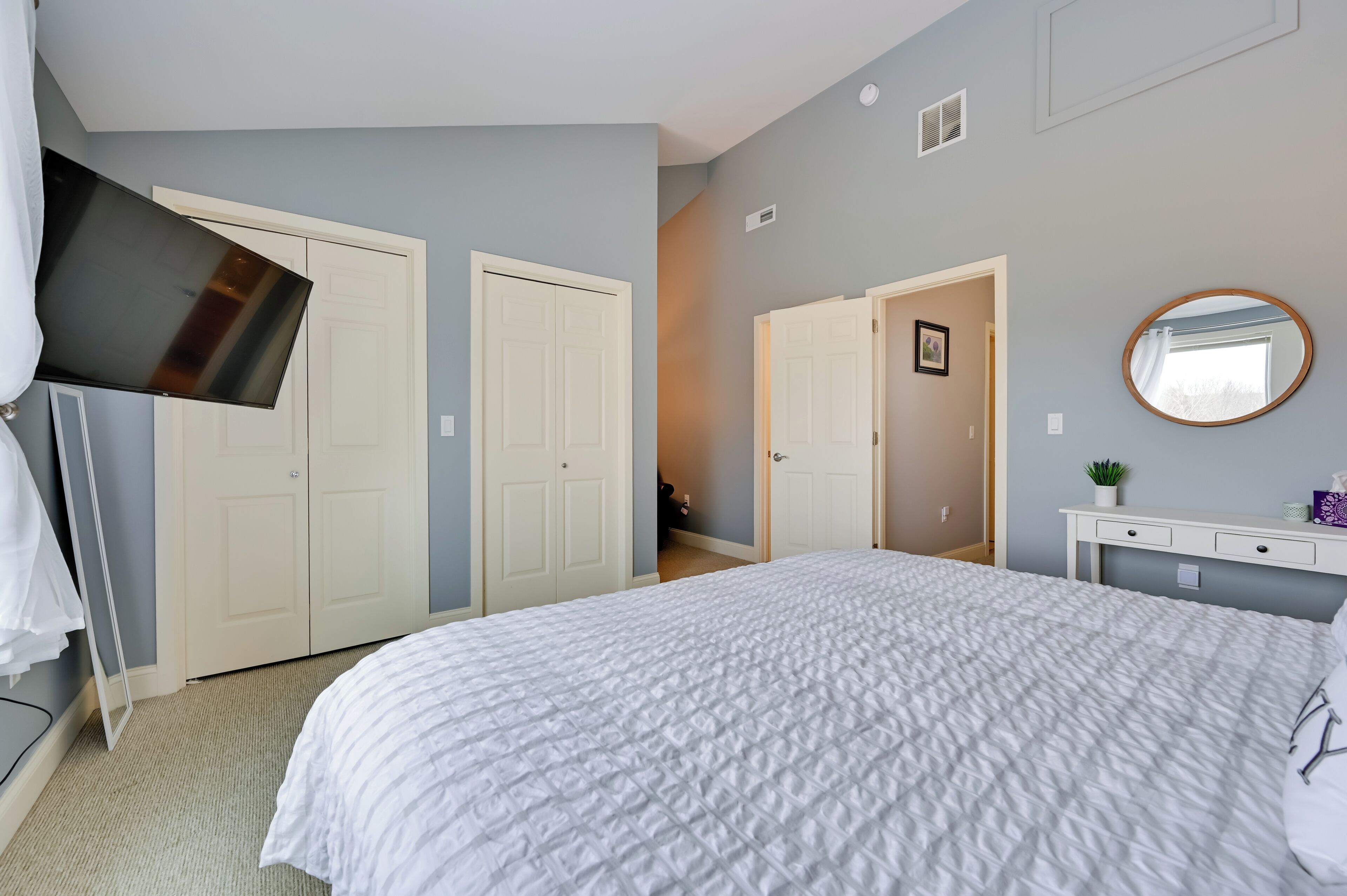 A bedroom with a large bed, two white doors, a flat-screen TV mounted on the wall, a small bedside table with a mirror, and a light-colored carpet.