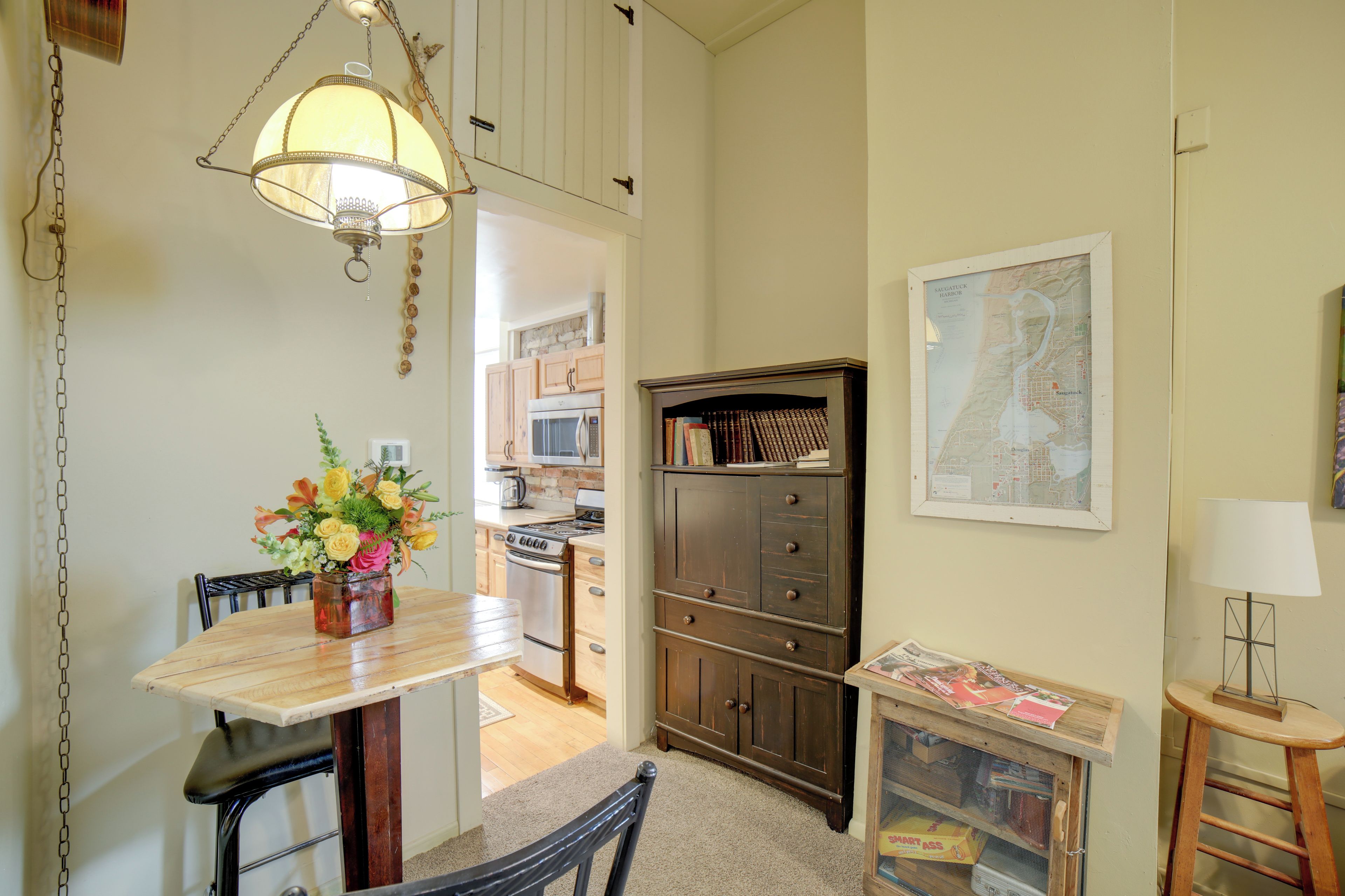 Un comedor con mesa de madera, sillas y una lámpara colgante. Se observa un mueble con libros y un mapa en la pared.
