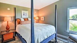 A four-poster bed with a white bedspread, a nightstand with a lamp, and a window with a view of greenery.