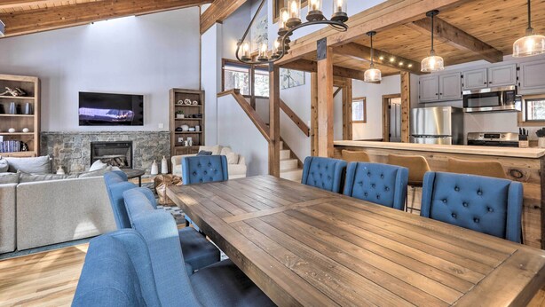 Un vaste espace de vie comprenant une grande table à manger en bois, des chaises recouvertes de tissu bleu, une cheminée en pierre et une cuisine équipée d’appareils électroménagers modernes.