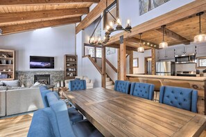 Een ruime woonkamer met een grote houten eettafel, blauwe gestoffeerde stoelen, een stenen openhaard en een keuken met moderne apparatuur.
