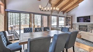 Une pièce spacieuse avec une grande table à manger en bois, entourée de chaises rembourrées, une cheminée et de grandes fenêtres donnant sur l’extérieur.