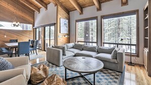 Un salon spacieux, doté d’un grand canapé d’angle, d’une table basse ronde et de grandes fenêtres offrant une vue sur la forêt.