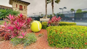 Een tennisbaan met een groot beeld van een tennisbal, omgeven door goed onderhouden hagen en palmbomen.