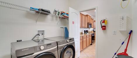 A laundry room with two Samsung washing machines, a shelf with folded clothes, a mop, and a fire extinguisher.