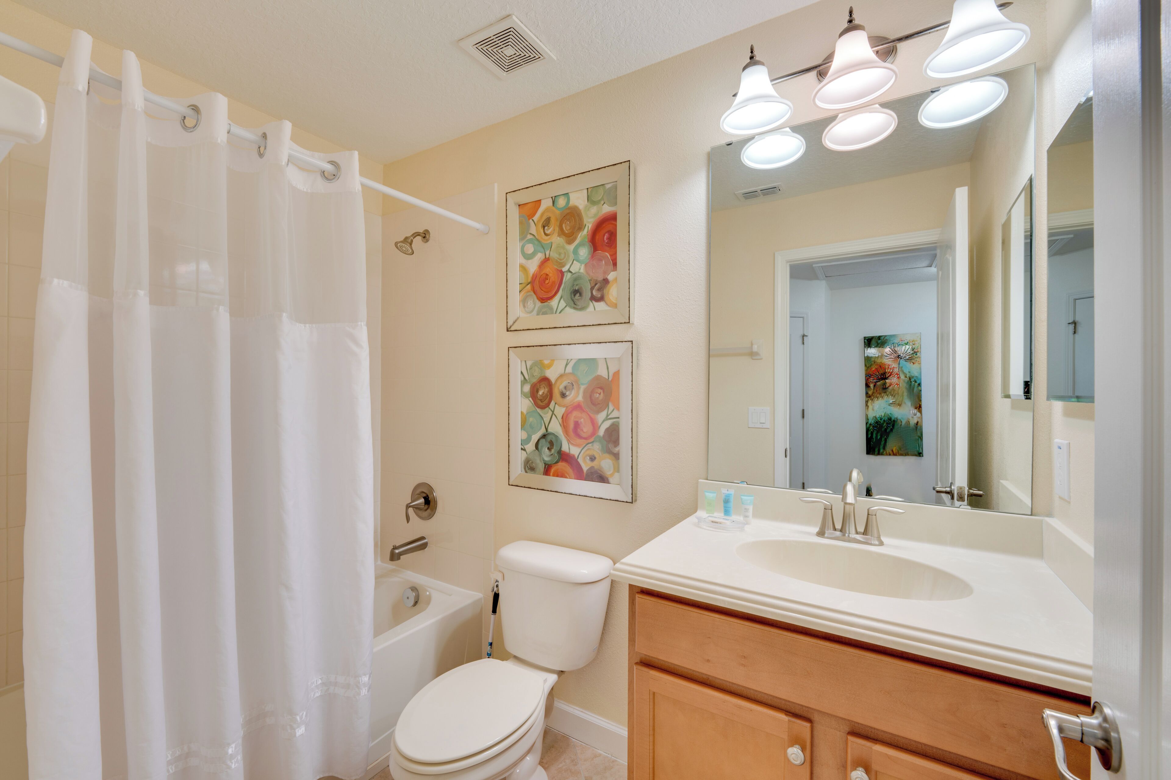 Une salle de bain avec une baignoire, des toilettes et un meuble-lavabo avec un miroir. Deux tableaux représentant des motifs floraux sont accrochés au mur.